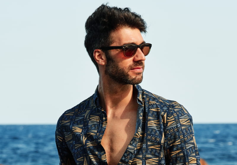 óculos de sol harmoniza o look tropical  jovem homem olhando pro lado com óculos de sol ao pro do sol na praia