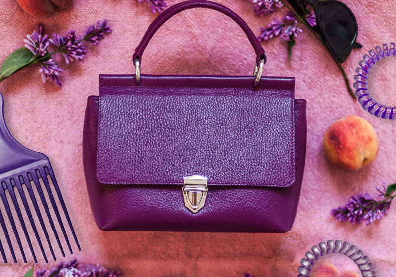 vista de cima de uma bolça lilás, óculos preto, prendedor de cabelo e pente afro lilás em cima de um lençol lilás rosado 