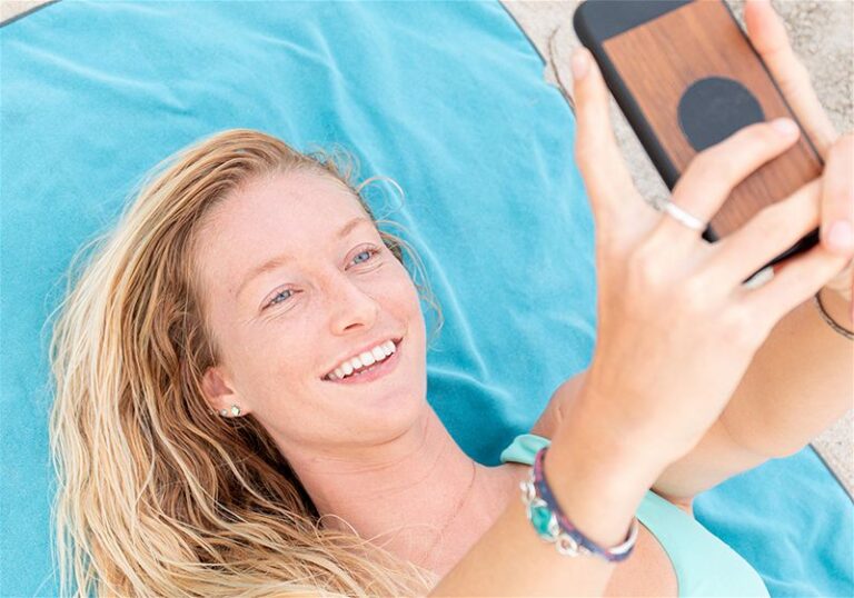 Mulher sorrindo deitada em cima da canga na praia e com e celular em cima tirando foto