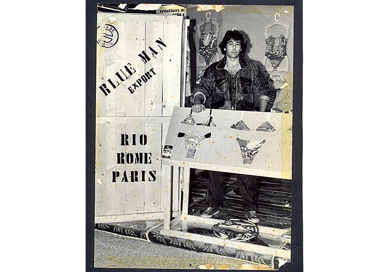 David Azulay dentro de espaço com biquínis em exposição, ao seu lado uma porta de madeira com a escrita Blueman e em sua frente um expositor de madeira com dois modelos de biquíni