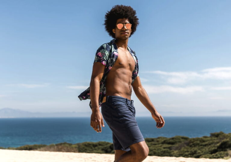 homem negro estilo andando na praia de camisa aberta, óculos de sol e bermuda