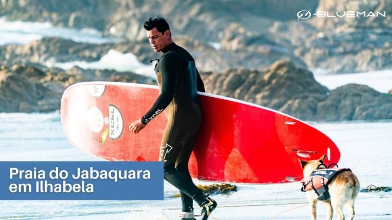 praia-do-jabaquara-em-ilhabela surfista segurando prancha vermelha com cão atrás de colete e mar ao fundo